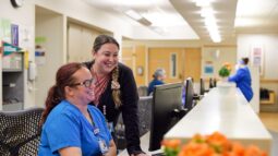 Two nurses smile at a computer at a nurses station.