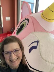 Woman in glasses smiles for camera with large inflatable pink unicorn