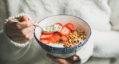 Healthy breakfast greek yogurt, granola and strawberry bowl in hands of woman wearing white loose knitted woolen sweater, selective focus. Clean eating, healthy, vegetarian, dieting food concept
