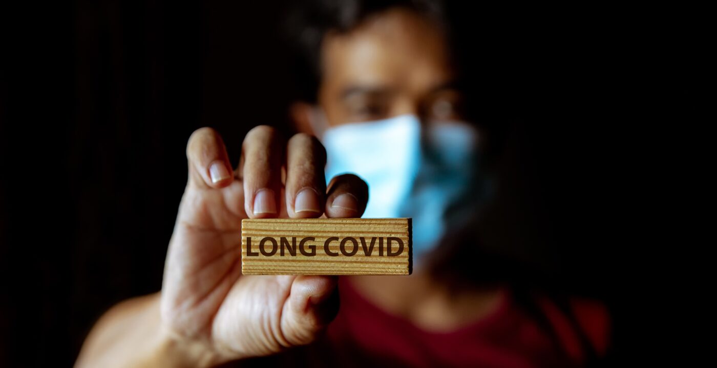 A masked person holding up a block that says long covid on it.