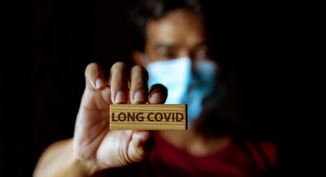 A masked person holding up a block that says long covid on it.