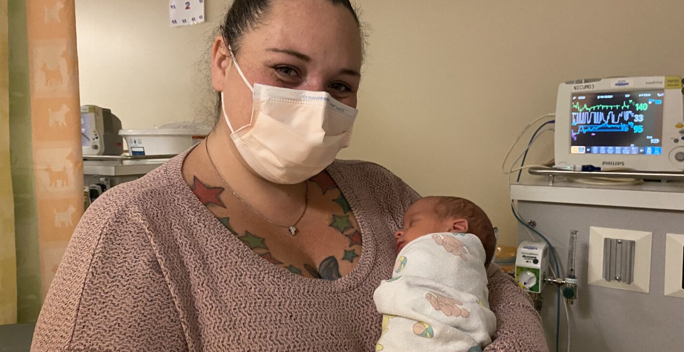 Woman in sweater holds swaddled infant in hospital room