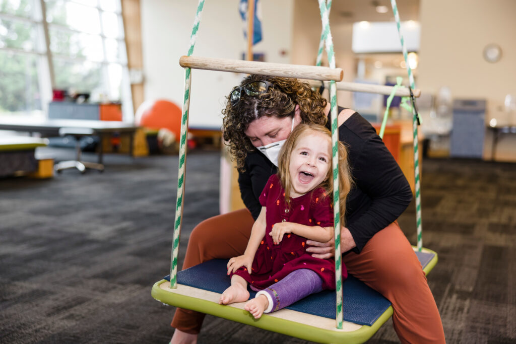 Smiling toddler wearing a red dress and purple orthotics on her left leg sits on a large swing with female physical therapist wearing a black top and orange pants