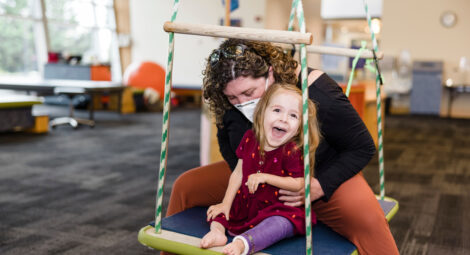 Smiling toddler wearing a red dress and purple orthotics on her left leg sits on a large swing with female physical therapist wearing a black top and orange pants