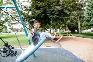 Smiling boy plays on swing at playground, while wheelchair sits in background