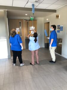 Three volunteers look up at a hospital ceiling while one uses a long duster to clean a light fixture