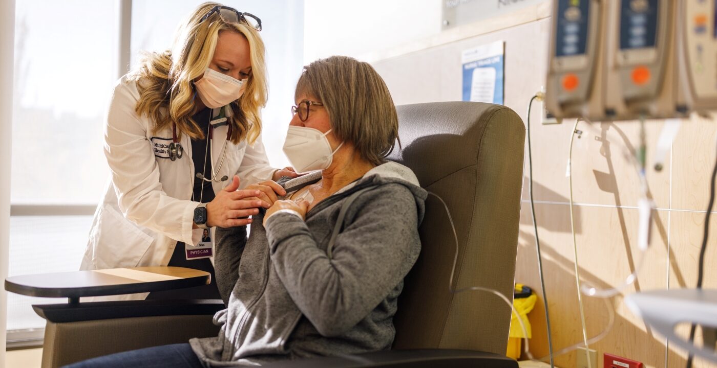 Doctor in white coat and face mask leans over patient to examine her picc line for cancer treatment