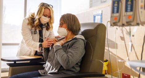 Doctor in white coat and face mask leans over patient to examine her picc line for cancer treatment