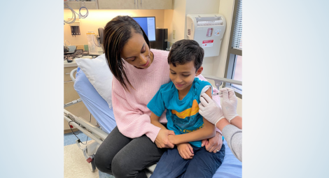 Woman in pink sweater sits next to child receiving a vaccine with one arm around his waist and the other holding his hand