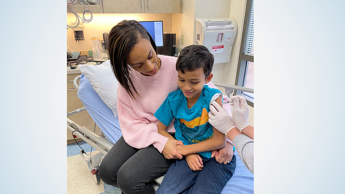 Woman in pink sweater sits next to child receiving a vaccine with one arm around his waist and the other holding his hand