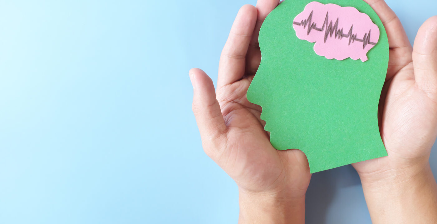Hands cradle a paper cutout of human head with pink brain showing heartbeat rhythm graph