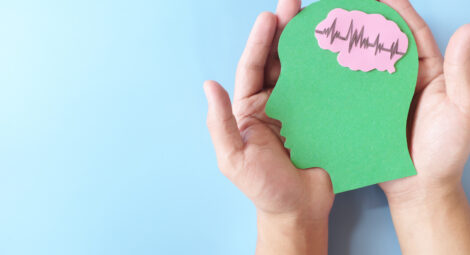 Hands cradle a paper cutout of human head with pink brain showing heartbeat rhythm graph