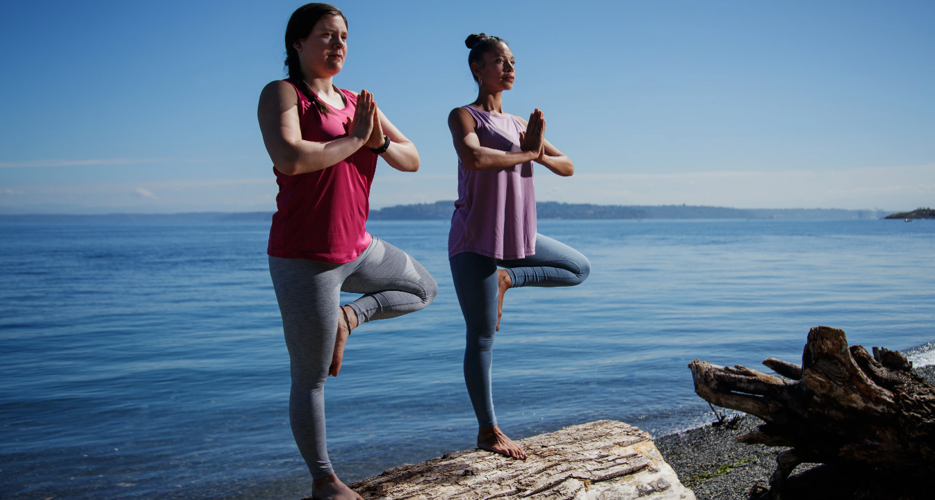 Two women doing yoga on a log by the water