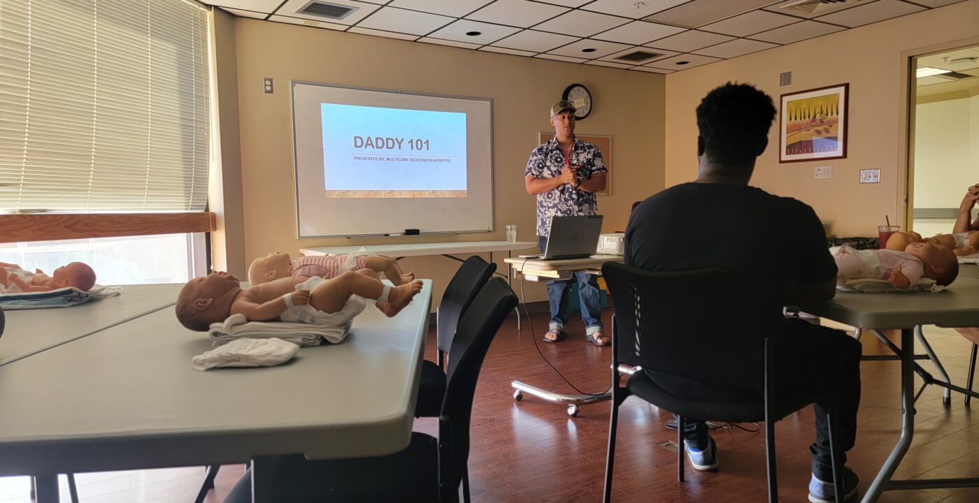 Man stands at front of classroom, “Daddy 101” presentation displayed on screen behind him; audience sits at tables with baby dolls