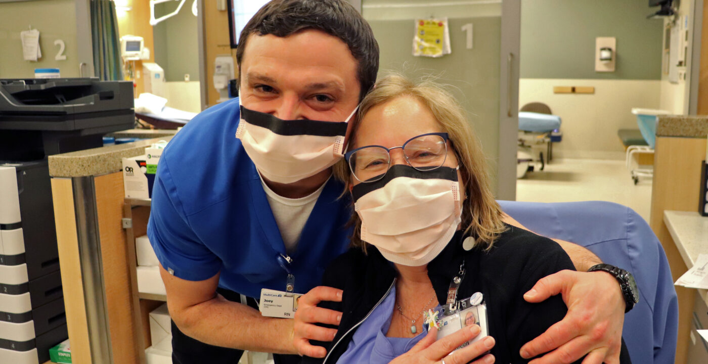 Two MultiCare employees embrace in an emergency department setting. Both are wearing paper masks.