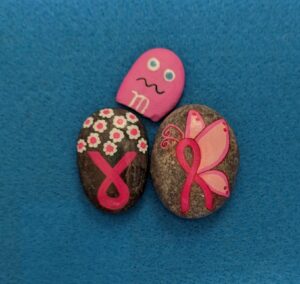 Three painted rocks: one painted like a pink M&M candy, one with a cancer ribbon and flowers and one with a butterfly