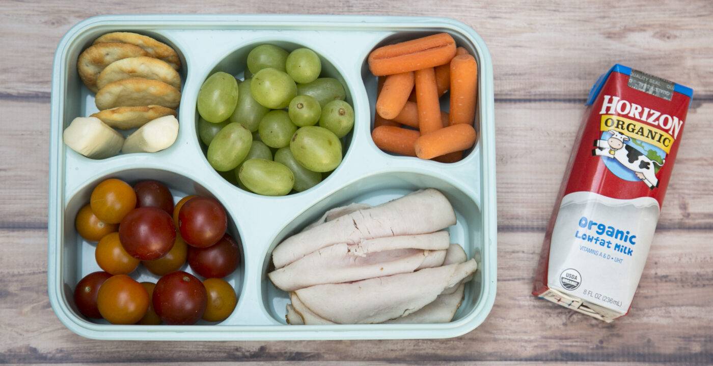 A bento box featuring rolled-up turkey slices, fruits, vegetables and a carton of milk.