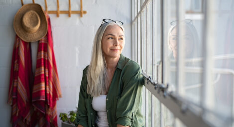 Smiling mature woman looking outside window. Thoughtful old woman looking away through window. Senior beautiful woman sitting at home with pensive expression.