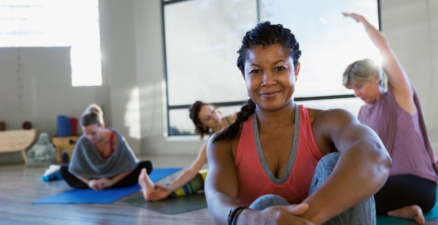 Woman smiles for camera while sitting on the floor in a bright room with lots of windows as other women stretch on mats behind her.