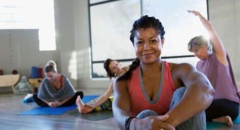 Woman smiles for camera while sitting on the floor in a bright room with lots of windows as other women stretch on mats behind her.