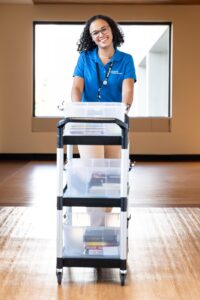 Woman in blue polo and khakis pushes a book cart down a hospital hallway