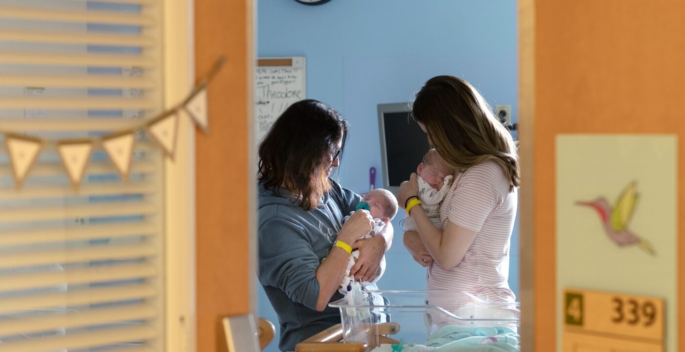 Two women are seen through an open door holding young infants in a hospital