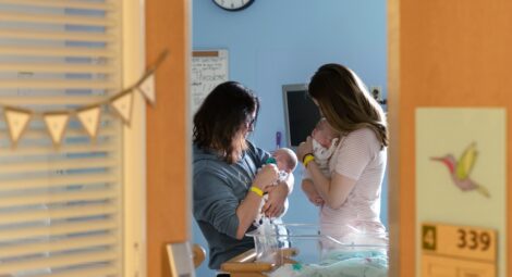 Two women are seen through an open door holding young infants in a hospital