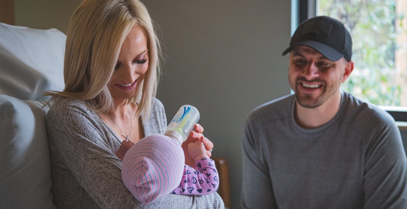 Mother feeding baby with a bottle while smiling man looks on