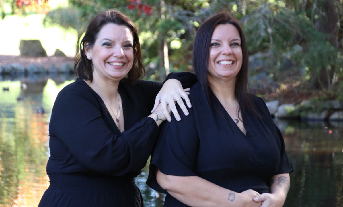 Twin women in black dresses and silver necklaces pose in front of a pond on a sunny day; one twin has short brown hair and the other has long dark brown hair