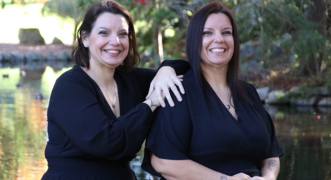 Twin women in black dresses and silver necklaces pose in front of a pond on a sunny day; one twin has short brown hair and the other has long dark brown hair