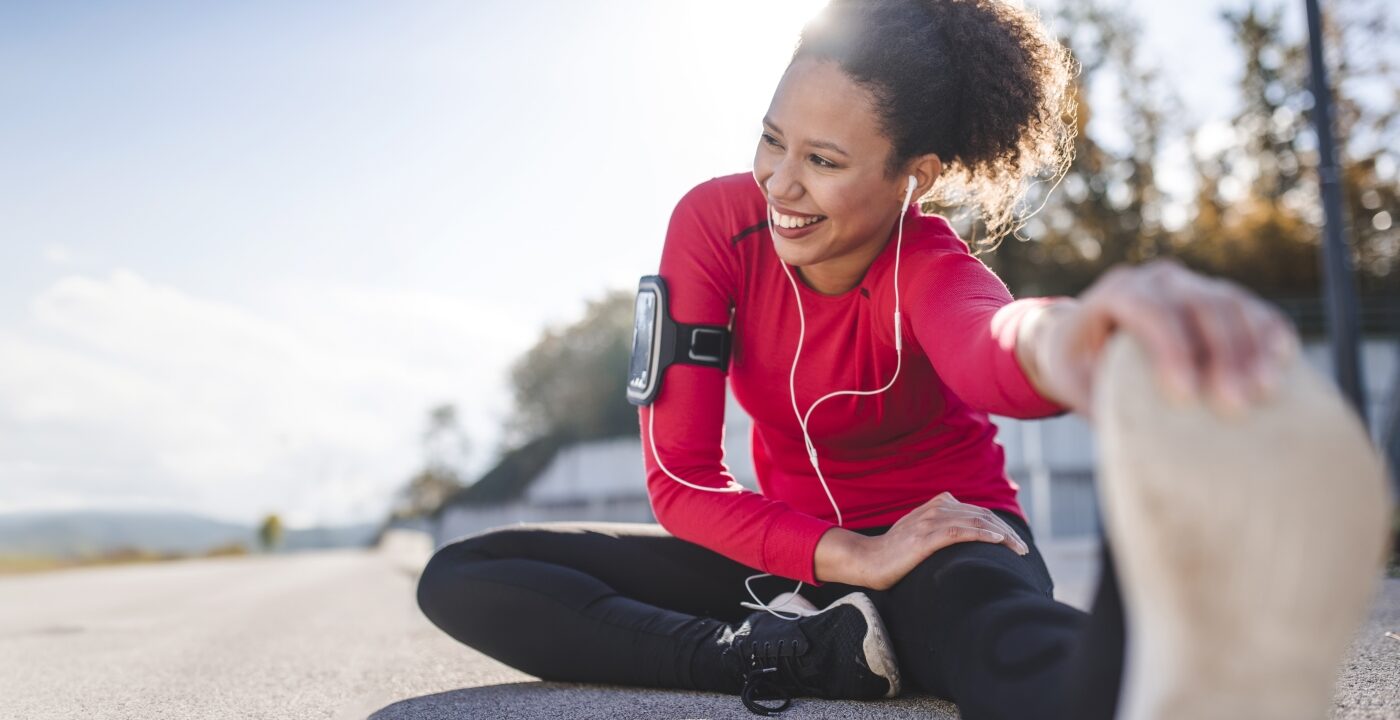 Woman in running gear sits on the pavement and stretches with a hand grabbing her shoe at the toes. It’s a sunny day and she has a big smile on her face.
