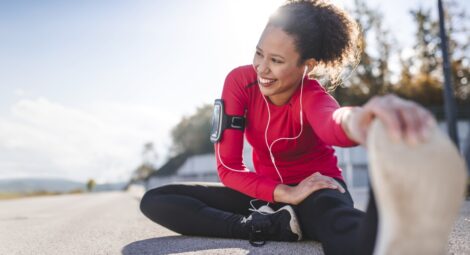 Woman in running gear sits on the pavement and stretches with a hand grabbing her shoe at the toes. It’s a sunny day and she has a big smile on her face.