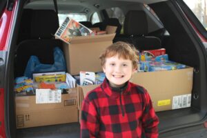 Boy in red plaid pullover smiles in front of a car trunk loaded with boxes of toys