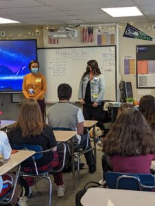 Two adults stand at the front of a classroom as students at desks listen