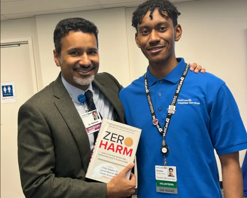 Man in suit holding “Zero Harm” book poses with young man in blue MultiCare Volunteer Services shirt