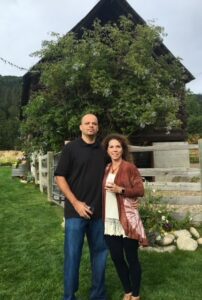 Man and woman standing in front of wooden fence and tree with wine glasses in hand