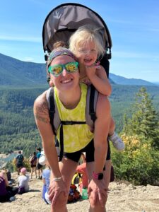 Woman in neon green tank top and sunglasses carrying a blonde girl on her back