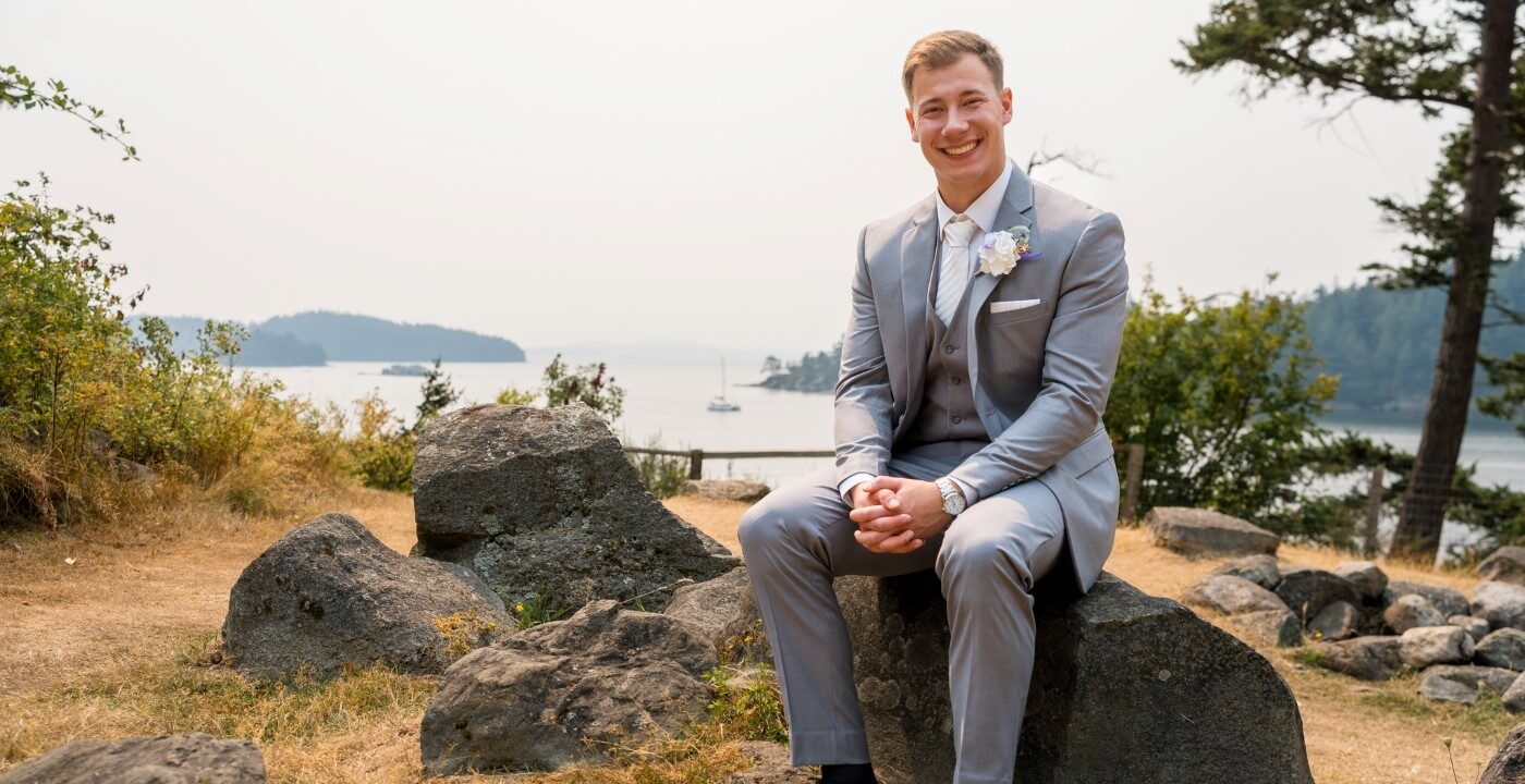 Young man wearing a gray suit sitting on a rock by the waterfront