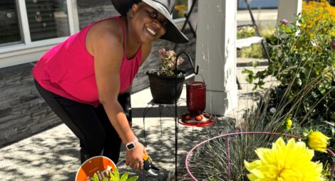 Woman in wide-brimmed hat holds plant clippers above a plant bed while smiling at the camera