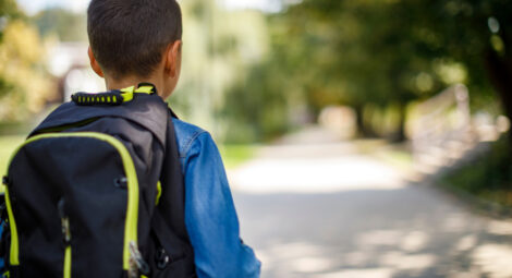 A child is seen from the back walking alone with a backpack on