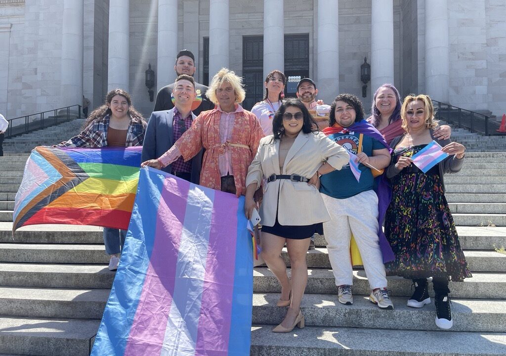 Group of people stand on concrete steps in the sun, some holding LGBTQ+ Pride flags
