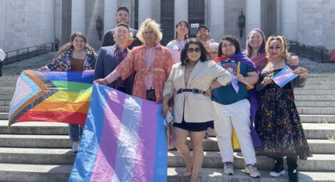 Group of people stand on concrete steps in the sun, some holding LGBTQ+ Pride flags
