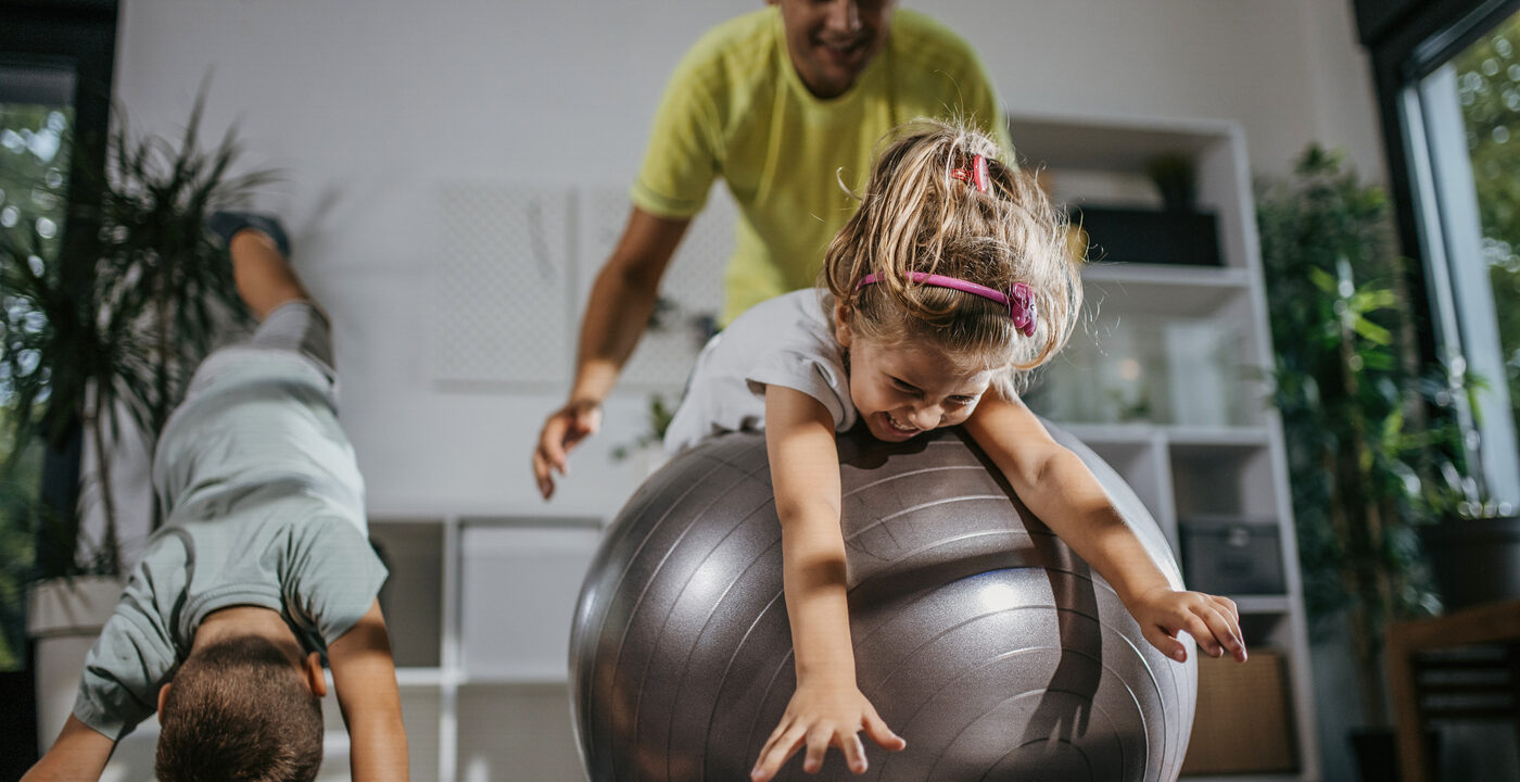 Child rolling on large workout ball with adult behind her and a young boy doing yoga next to them