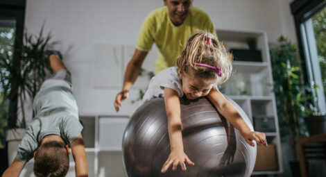 Child rolling on large workout ball with adult behind her and a young boy doing yoga next to them