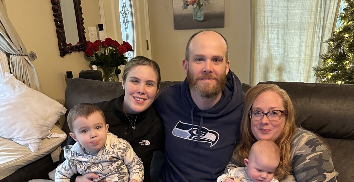 Two adult women, an adult man, a toddler and an infant pose for a family photo on a couch in a living room during Christmas