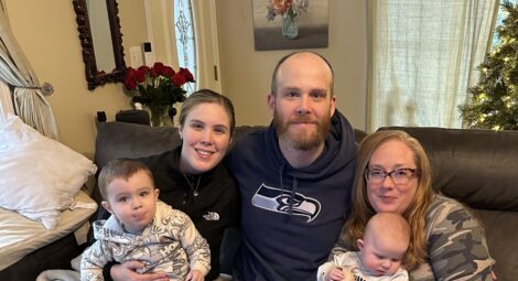 Two adult women, an adult man, a toddler and an infant pose for a family photo on a couch in a living room during Christmas