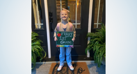 Young girl standing on doorstep holding a sign that says “first day of 1st grade”