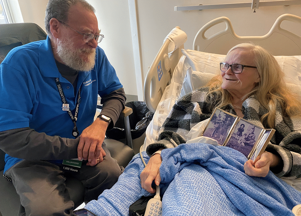 Man sits next to older woman in hospital bed who shows off framed photos