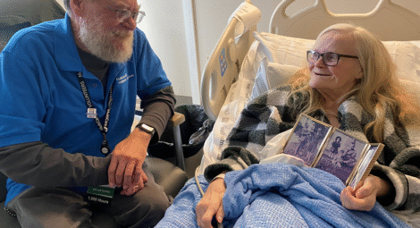 Man sits next to older woman in hospital bed who shows off framed photos