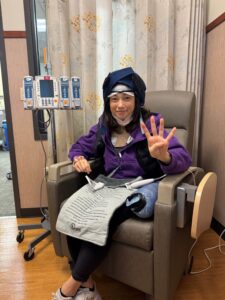 woman holds up four fingers while sitting in chair, Caption: Gabler at her fourth and final chemo treatment